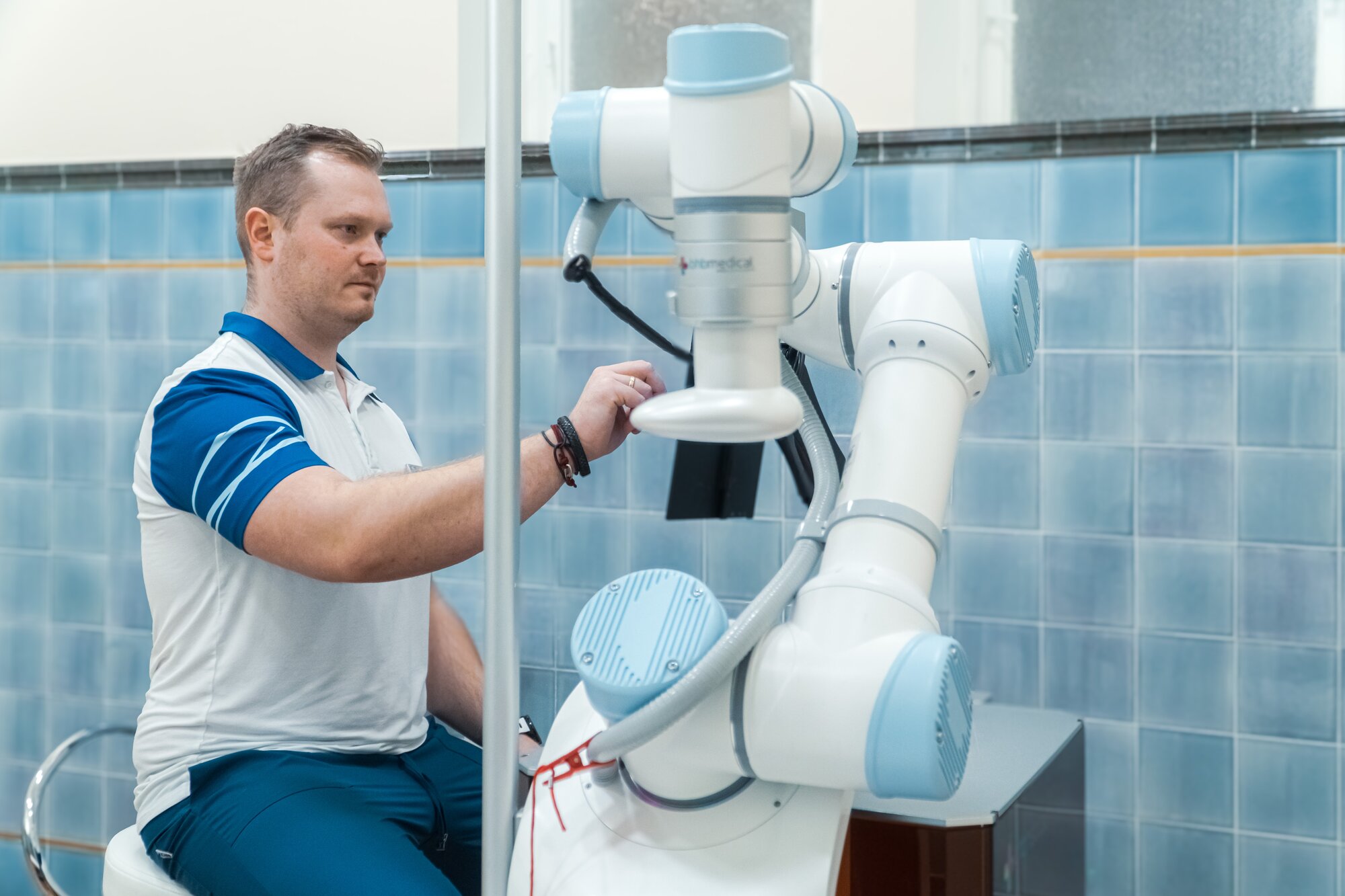 Physiotherapist at the robotic arm of the Rocozyer massage device in a treatment room with blue wall tiles