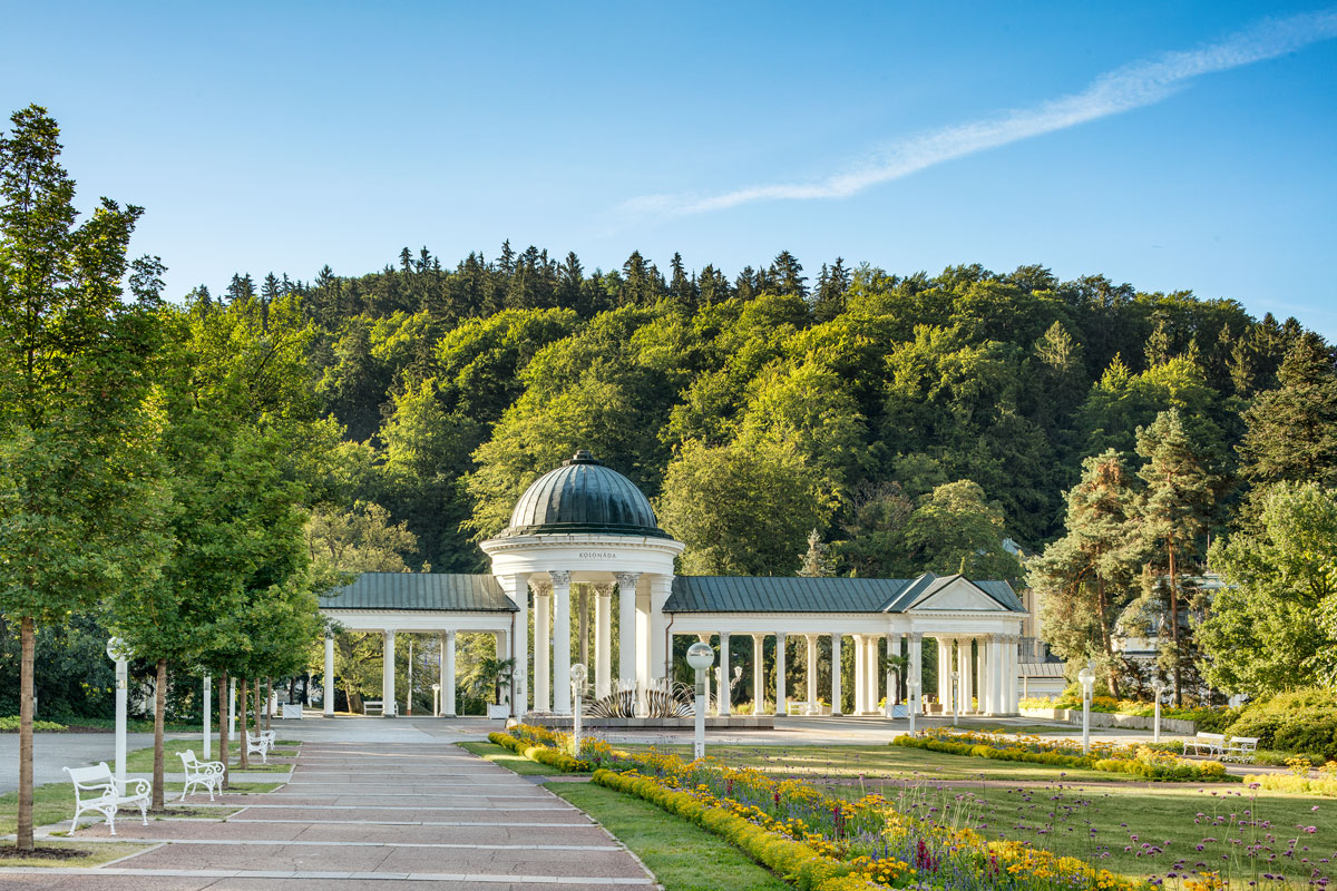 Weiße Säulenhalle der Karolinen- und Rudolfsquelle im Kurpark, dahinter ein bewaldeter Hang