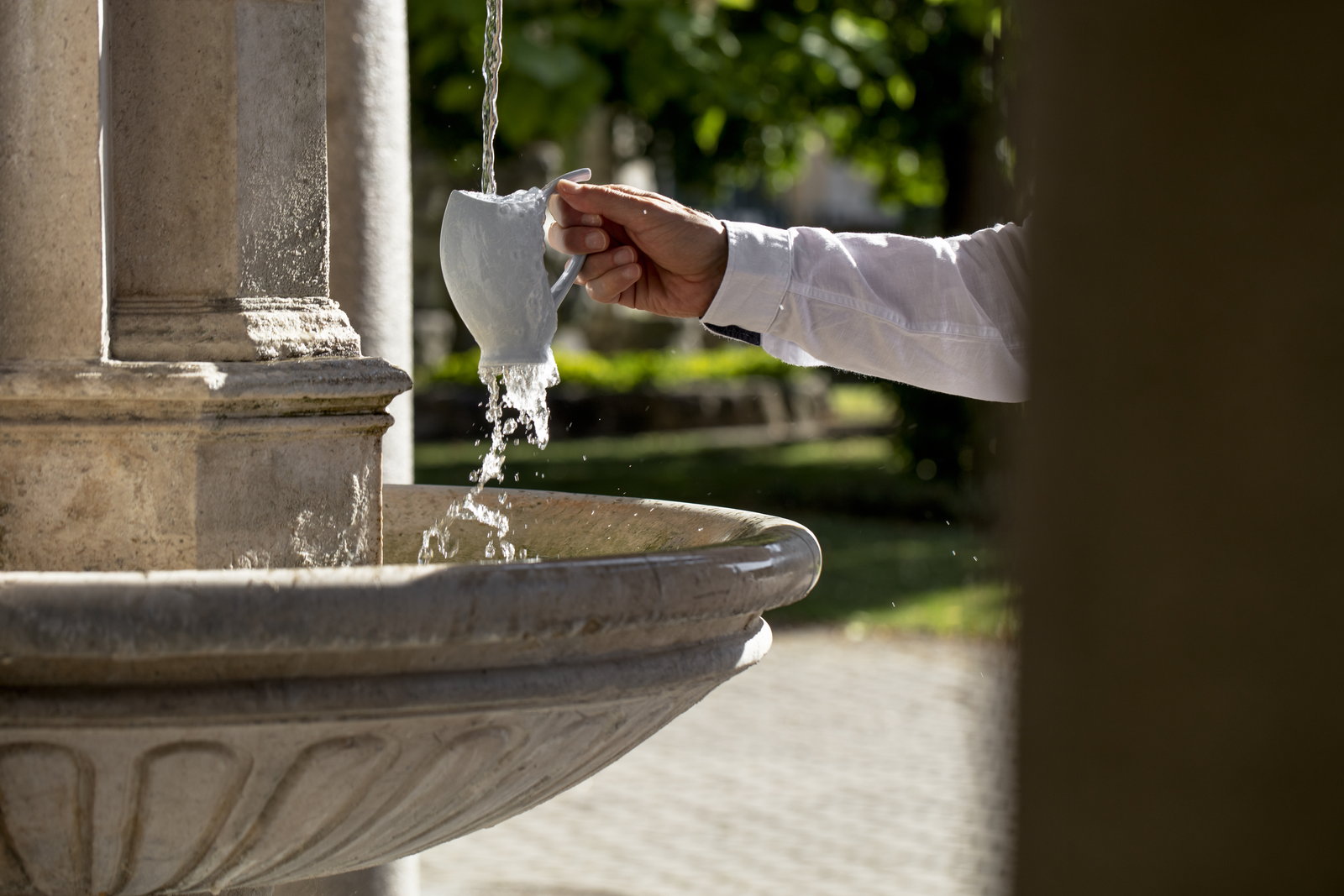 A hand holds a white porcelain spa cup under a stream of mineral water at a colonnade pillar