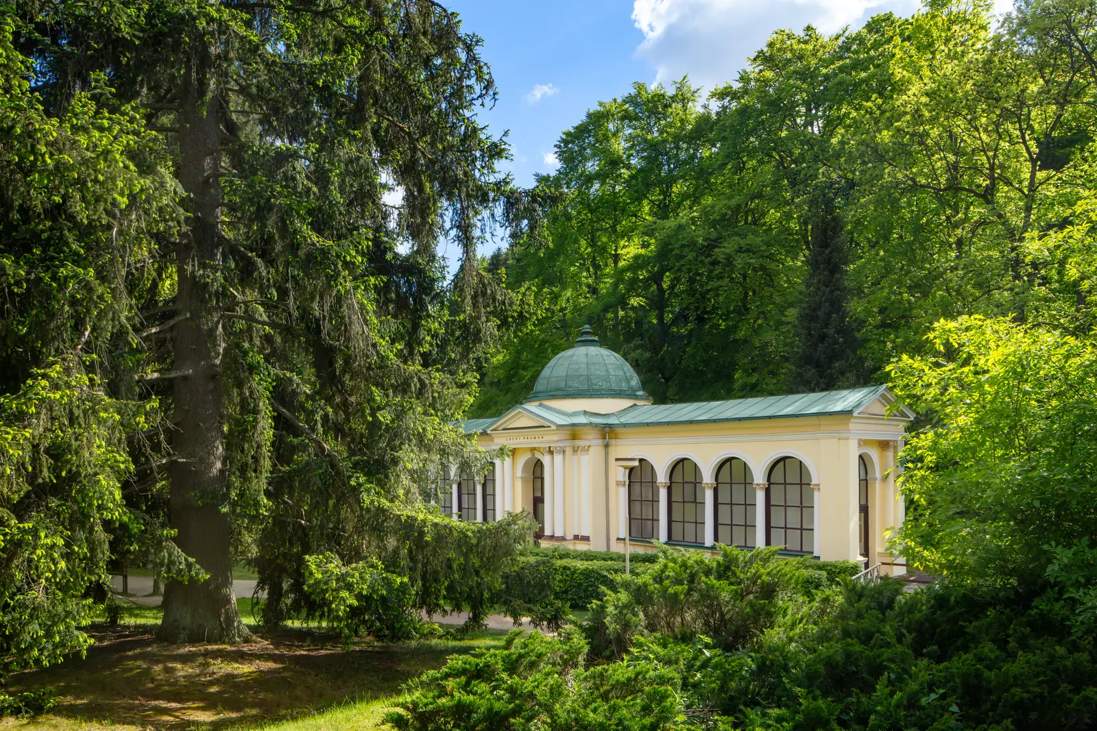 Pavillon der Waldquelle mit grüner Kuppel zwischen hohen Fichten und Laubbäumen