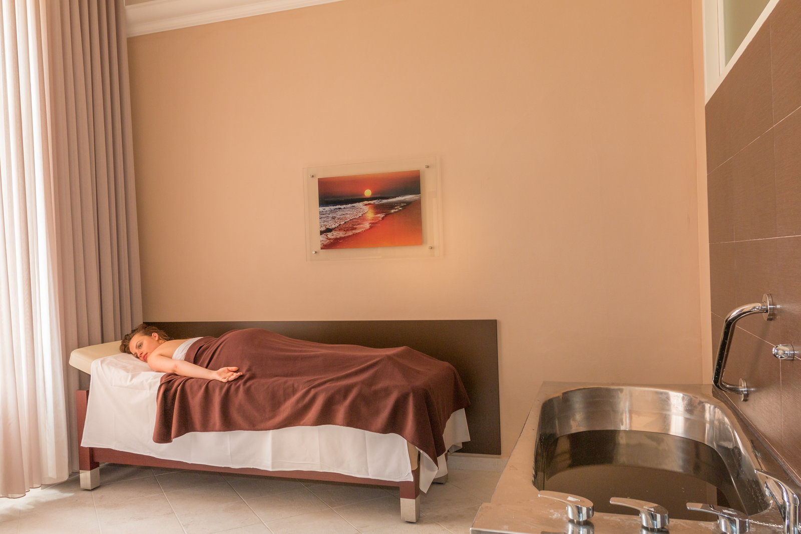 A woman rests on a treatment bed under a brown blanket after her mineral bath, with the empty tub beside her