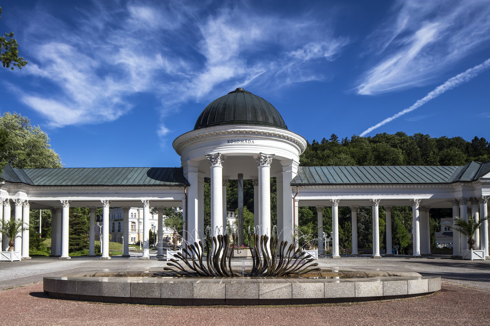 Domed white spring pavilion with colonnaded wings and a dark bronze fountain sculpture