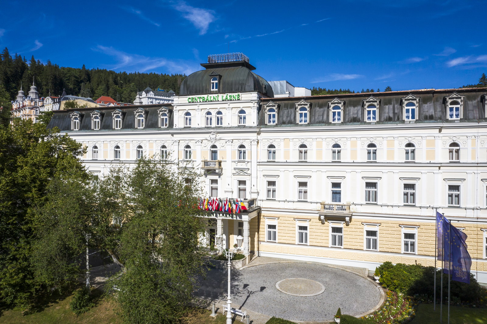 Aerial view of the Centrální lázně spa building and its domed tower below a wooded slope