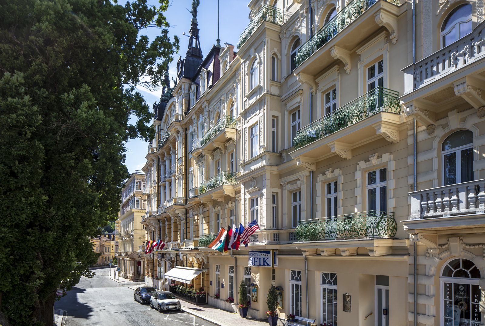 A row of ornate nineteenth-century spa facades with wrought-iron balconies above a tree-lined street