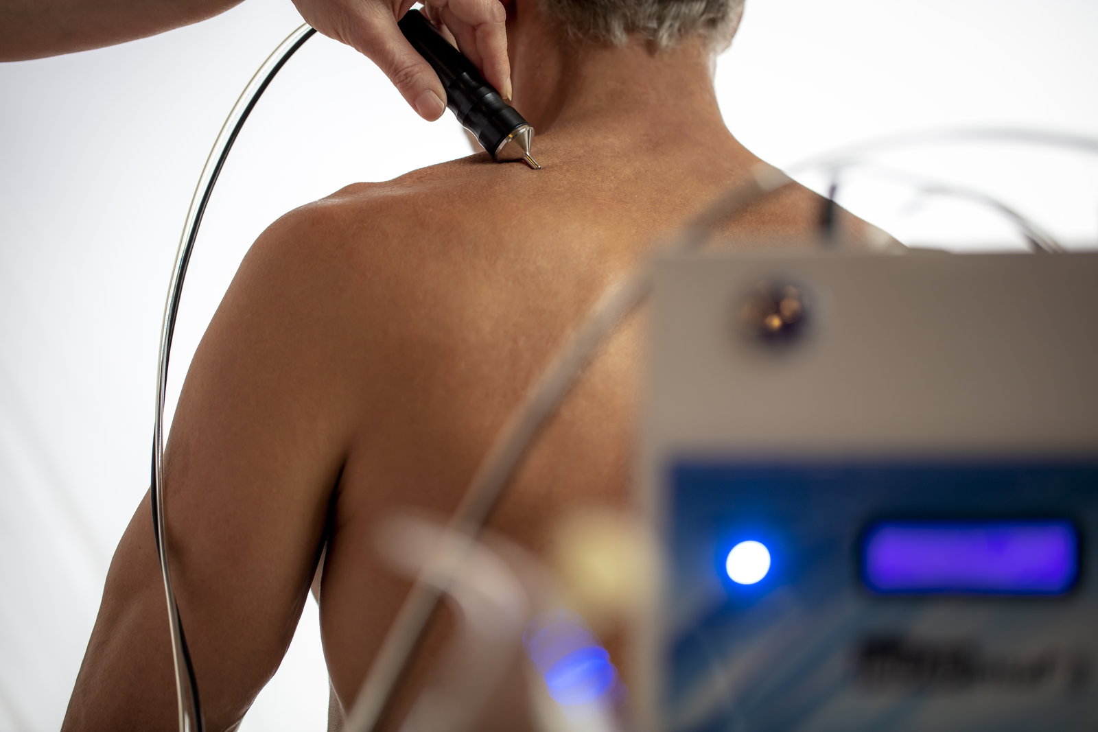A therapist holds a handheld probe to a patient’s neck and shoulder, connected to a device